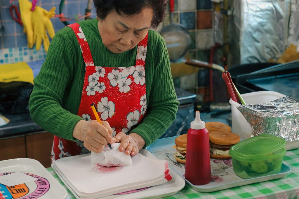 An elderly woman preparing food in a home kitchen, symbolizing food, habit, memory, and everyday life.